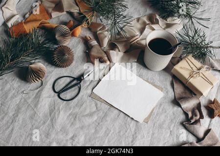 Natale ancora vita. Mockup biglietto d'auguri vuoto. Tazza di caffè, ornamenti di carta e confezione regalo. Nastri di velluto, rami di pino con forbici. Biancheria Foto Stock