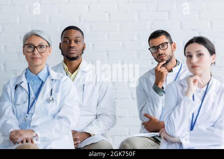 medici multietnici concentrati in cappotti bianchi seduti durante la conferenza Foto Stock