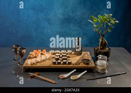 Ancora vita con rotoli di uramaki, maki di vari sapori, salmone e gamberi nigiri, ficus zenzero bonsai, salsa di soia e bacchette Foto Stock