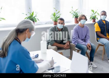 annebbiamento della scrittura dell'infermiera vicino a persone multietniche in maschere mediche in attesa nel centro di vaccinazione Foto Stock