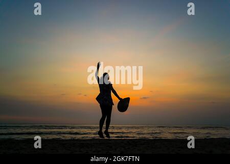 Vista posteriore della giovane donna isolata che salta in aria, tenendo un grande cappello da sole, al tramonto su una spiaggia del Regno Unito, vacanza estiva staycation. Salta per la gioia. Foto Stock