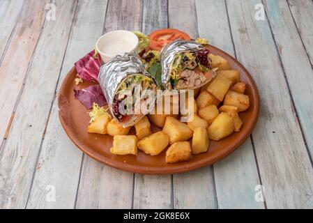 burrito con pollo e molte lattughe arrotolate con foglio di alluminio e una guarnitura di patatine fritte a dadini su un piatto di argilla Foto Stock