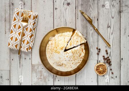 Immagine dall'alto della torta di carote con panca ghiacciata, piatti dorati e posate su tavola bianca di legno con chicchi di caffè e arancio secco Foto Stock