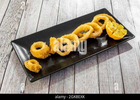Tapa di calamari in pastella affettata ad anelli su un piatto nero con limone affettato Foto Stock