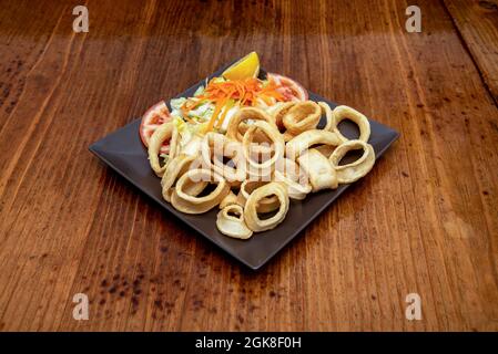 Piatto nero quadrato con coperchio spagnolo di anelli di calamari di pastella romana su tavolo di legno scuro Foto Stock