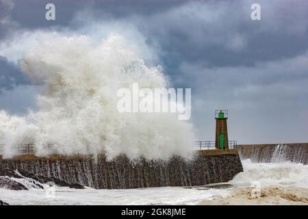 Enormi onde di mare schiumose che si infrangono contro il frangiflutti di pietra con la vecchia torre faro contro il cielo nuvoloso blu nel Porto di Viavelez nelle Asturie Spagna Foto Stock