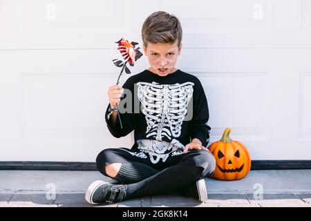 Corpo pieno di ragazzo preteen creepy in costume scheletro nero con il trucco tenendo il fiore e guardando la macchina fotografica mentre si siede vicino lanterna di zucca contro Foto Stock