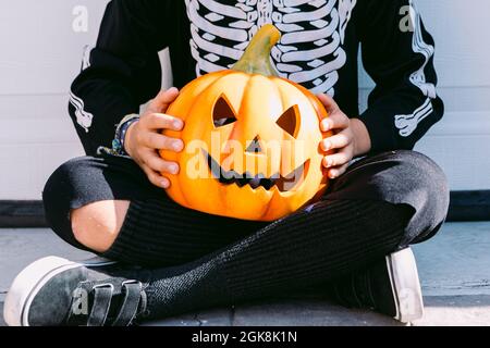 Raccolto irriconoscibile bambino in nero scheletro costume che tiene creepy scolpito Halloween Jack o Lantern zucca mentre seduto con le gambe incrociate sulla strada Foto Stock