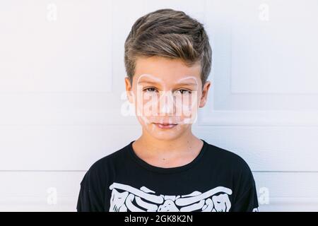 Ragazzo serio preda con trucco scheletro dipinto di bianco vestito in costume nero di Halloween guardando la macchina fotografica contro la parete bianca Foto Stock