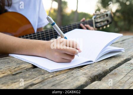 Adolescente femminile ritagliato e irriconoscibile con chitarra classica che suona corda mentre scrive musica in copybook nel parco su sfondo sfocato Foto Stock