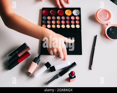 Alto angolo di raccolto anonimo capretto con eyeshadow e tavolozza di glitter tra specchio e vari prodotti di bellezza in casa Foto Stock