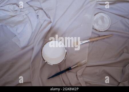 Vista dall'alto del contenitore di vernice bianca con coperchio e spazzole su panno sgualcito durante la ristrutturazione in casa Foto Stock