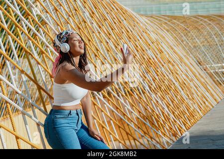 Positiva giovane etnia femminile con cellulare che prende un selfie mentre ascolta la musica dalle cuffie wireless mentre ride in città Foto Stock