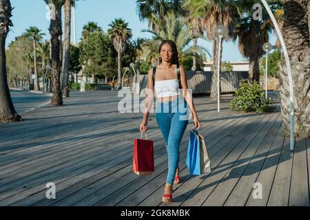 Allegro giovane acquirente afroamericano femmina con shopping borse guardare via mentre si cammina per strada Foto Stock