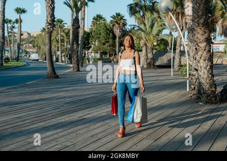 Allegro giovane acquirente afroamericano femmina con shopping borse guardare via mentre si cammina per strada Foto Stock