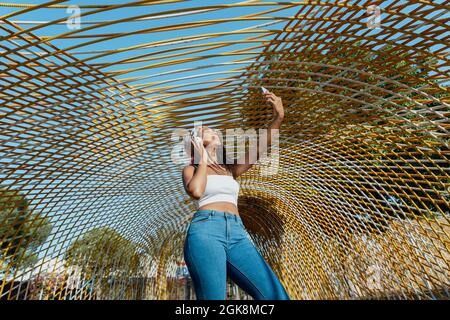 Positiva giovane etnia femminile con cellulare che prende un selfie mentre ascolta la musica dalle cuffie wireless mentre ride in città Foto Stock