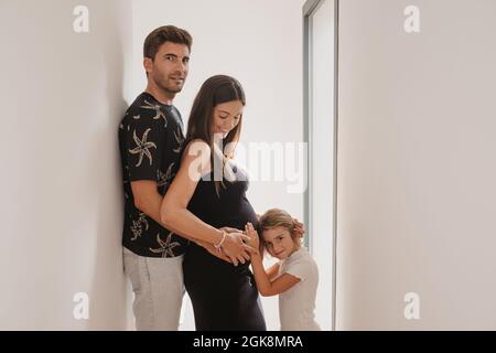 Vista laterale del bambino carezzando il ventre della madre incinta sorridente contro il papà mentre nel passaggio a casa Foto Stock