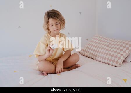 Bambino a piedi nudi in t shirt con succhietto seduto con gambe incrociate su un soffice letto in casa Foto Stock