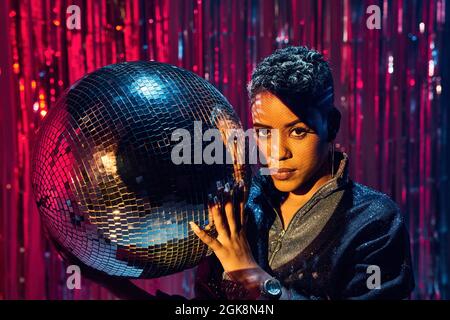 Giovane etnia trendy che guarda la macchina fotografica mentre tiene la palla a specchio nel nightclub Foto Stock