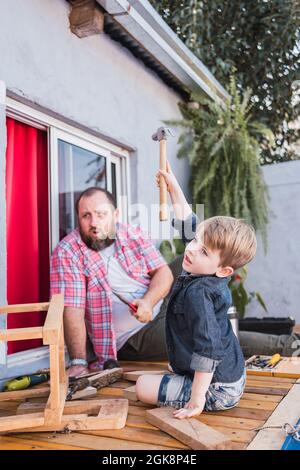 Papà bearded che insegna figlio con martello che lavora con legno mentre si siede sul lungomare il fine settimana Foto Stock