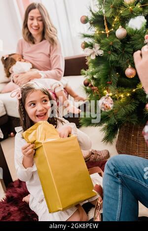 Bambino sorridente che apre la scatola presente tra il padre del raccolto e la madre che allattano il bambino durante la festa di Capodanno a casa Foto Stock