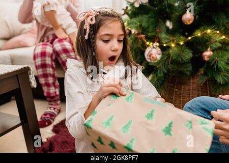 Sorprende bambino che apre la scatola presente fra i genitori del raccolto ed il fratello mentre festeggia la festa di Capodanno in casa Foto Stock