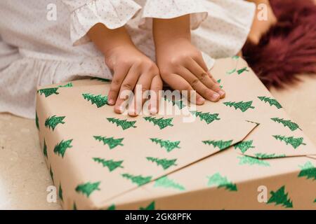 Crop anonymous bambino apertura presente scatola con abete modello sul pavimento durante Capodanno vacanza in casa Foto Stock