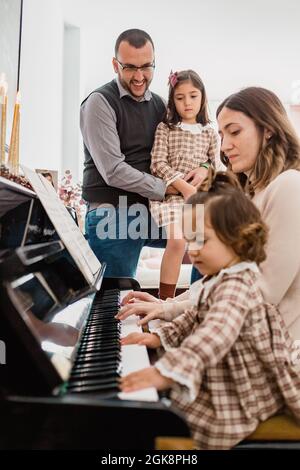 Adorabile bambino toddler con mamma che suona il pianoforte contro il padre sorridente che abbraccia la figlia nella stanza della casa Foto Stock