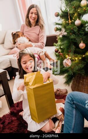 Bambino sorridente che apre la scatola presente tra il padre del raccolto e la madre che allattano il bambino durante la festa di Capodanno a casa Foto Stock