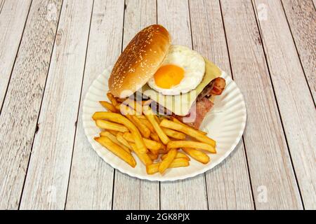 Hamburger di manzo con uova fritte, formaggio affettato, pancetta fritta, sottaceti e lattuga con porzione di patatine fritte su piatto bianco Foto Stock