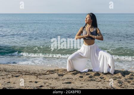 Ragazza asiatica sognante con le mani in preghiera che squattano in posizione Utkata Konasana mentre praticano lo yoga e si guarda via sulla riva del mare Foto Stock