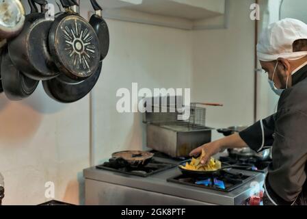 Vista laterale del raccolto etnia maschio cuoco in maschera uniforme e sterile preparazione della pasta in padella in cucina ristorante Foto Stock