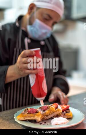 Raccolto etnico maschio cuoco in maschera sterile decorando deliziosi waffle viennesi con panna montata da bottiglia in cucina ristorante Foto Stock