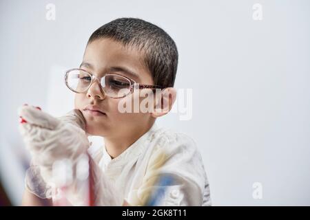 Un ritratto di un ragazzo carino in guanti e occhiali protettivi che tengono un pezzo di plastilina rossa in mani contro lo sfondo bianco Foto Stock