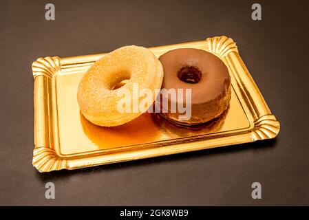 Vassoio d'oro con un paio di ciambelle di cioccolato e zucchero con i loro fori su un tavolo scuro Foto Stock