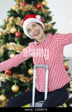 Donna sorridente in cappello babbo natale siede sulla valigia sullo sfondo dell'albero di Natale Foto Stock