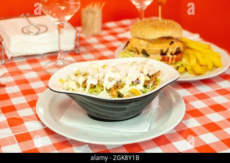 Insalata Caesar con pollo, formaggio affettato, uova sode, salsa di maionese, lattuga iceberg su una ciotola da lavoro e una tovaglia rosso chillon ristorante Foto Stock