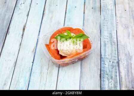 Palla di burrata fresca con fette di pomodoro, pepe e basilico all'interno di un pacchetto di consegna a domicilio su sfondo di legno Foto Stock