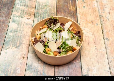 Insalata Caesar con scaglie di parmigiano, crostini e germogli di lattuga con rucola in recipiente di cartone per la consegna a domicilio su tavola di legno Foto Stock