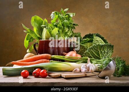 Verdure fresche assortite con tagliere, mestolo e vecchio crock pot in ambiente rustico su tavola di legno e sfondo giallo. Foto Stock