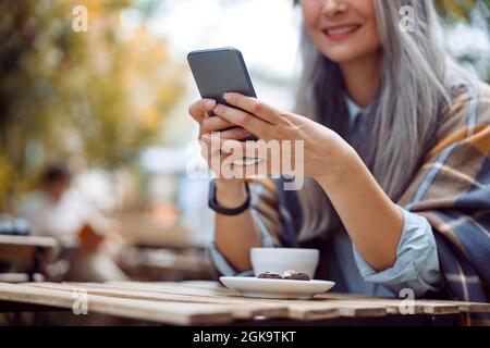 Signora asiatica matura felice scrive il messaggio sul telefono seduto al tavolo sulla terrazza del caffè all'aperto Foto Stock