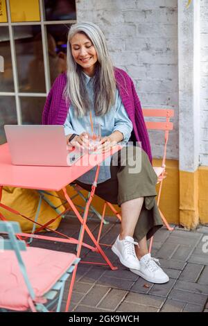 Sorridente donna asiatica con vetro d'acqua siede al tavolo di corallo con un laptop aperto Foto Stock