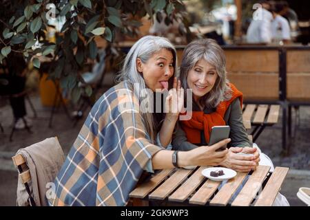 Donna asiatica matura grimaces e amico con capelli d'argento a videochat in Street cafe Foto Stock