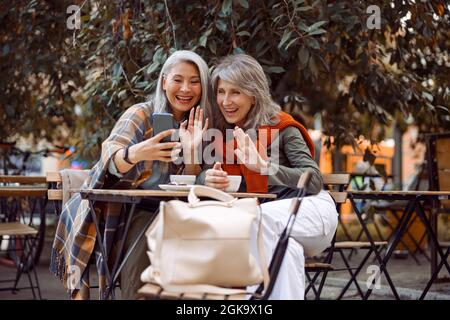 Buone donne maturo amici agitare le mani alla videochat via smartphone in Street cafe Foto Stock