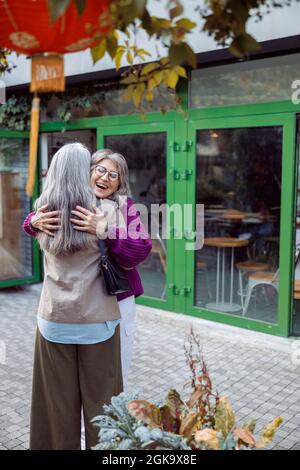 Signora anziana gioiosa in giacca calda abbracci lungo capelli migliore riunione amico sulla strada della città Foto Stock