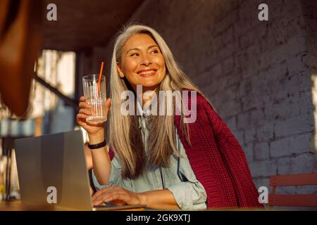 Una donna asiatica piuttosto anziana lavora su un moderno computer portatile al tavolo sulla terrazza del caffè all'aperto Foto Stock