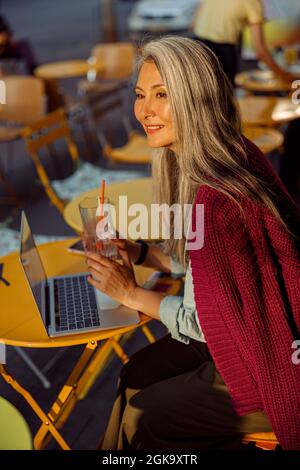 La signora asiatica mature e premurosa tiene il bicchiere con una bevanda seduta a un piccolo tavolo con un computer portatile in un bar Foto Stock