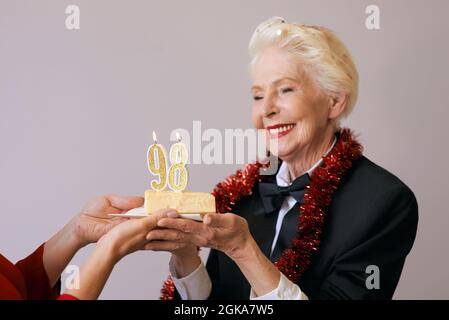 Felice allegra elegante novantotto anni donna in abito nero che celebra il suo compleanno con torta. Stile di vita, positivo, moda, stile concetto Foto Stock