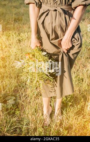 Una donna in abito verde con un bouquet di fiori selvatici si erge sull'erba senza scarpe. Le mani femminili che tengono un bouquet di fiori selvatici. Giorno d'estate. Foto Stock