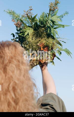 Bella donna che tiene un bouquet di fiori selvatici. Una bella donna bionda di mezza età in un abito verde con capelli ricci. Sfondo cielo blu estivo. REA Foto Stock